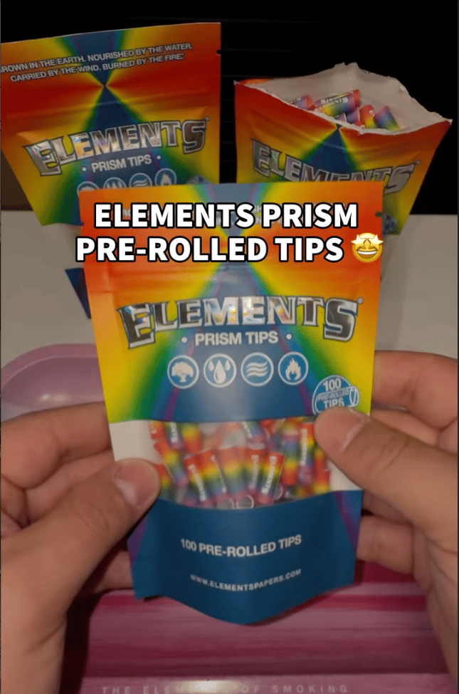 Two hands holding a colorful pouch of Elements Prism Tips with a clear window showing rainbow-colored pre-rolled tips inside. A text overlay reads 'ELEMENTS PRISM PRE-ROLLED TIPS' with a starry-eyed emoji, and additional pouches are visible in the background